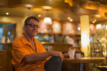 Obraz premium Man holding mug of coffee with a smile enjoying life in a Cafe wearing a orange shirt