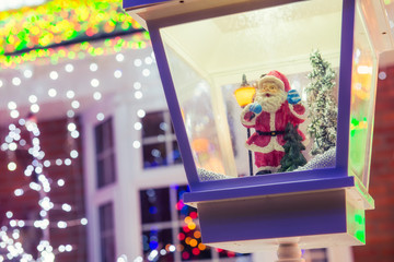 Close up Santa Claus in decorative lantern in the snow on the blurred Christmas Lights Background and outdoor decorations. Merry Xmas and Happy New Year concept. Selective focus, copy space.