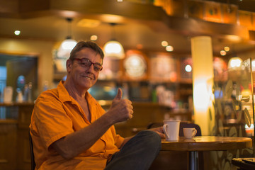 Man holding mug of coffee with a smile and thump up enjoying successful life  in a Cafe wearing a orange shirt