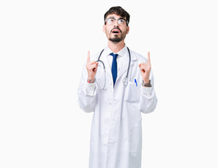 Fototapeta premium Young doctor man wearing hospital coat over isolated background amazed and surprised looking up and pointing with fingers and raised arms.