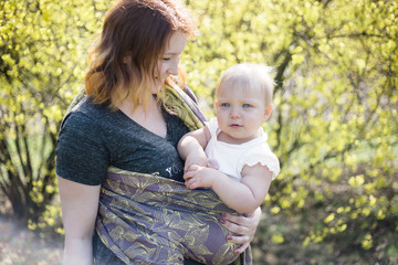 Fototapeta premium Mother holding her daughter wrapped in a purple sling, looking at her lovingly. Sunny, lifestyle, high key capture.