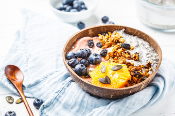 Breakfast smoothie bowl with chia pudding, berries and granola in a coconut shell on white wooden background.