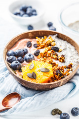 Breakfast smoothie bowl with chia pudding, berries and granola in a coconut shell on white wooden background.