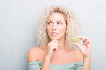 Young blonde woman over grunge grey background eating macaron serious face thinking about question,...