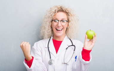 Young blonde dietitian doctor woman over grunge grey wall holding green apple screaming proud and celebrating victory and success very excited, cheering emotion