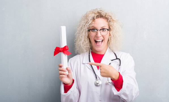 Young blonde doctor woman over grunge grey wall holding degree very happy pointing with hand and finger