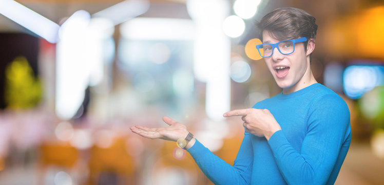 Young handsome man wearing blue glasses over isolated background amazed and smiling to the camera while presenting with hand and pointing with finger.