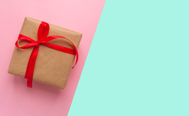 Gift box wrapped in brown colored craft paper and tied with red bow on pink and blue background with copyspace.