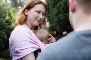 Woman carrying a baby in a pink sling, outdoors, looking at her anonymous partner.  Lifestyle, shallow depth of field.