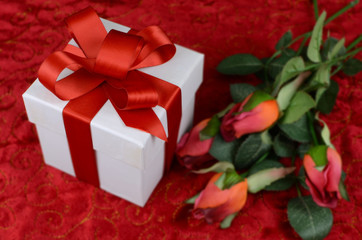 Beautiful image in shades of red for Valentine's Day in February. Silk roses and gift on velvety table cloth. Selective focus on bow