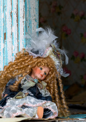 An old doll sitting against the door frame in an abandoned house. Debris litters the floor, paint is peeling from the frame. Portrait orientation.