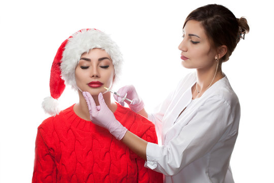 The doctor cosmetologist makes injection on the lips of a beautiful woman in the Santa Claus hat. New Year's and Cosmetology concept
