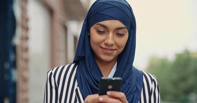 Young muslim girl wearing hijab walking down the street, texting someone on phone or surfing the web and expressively smiling - modern muslim concept 4k
