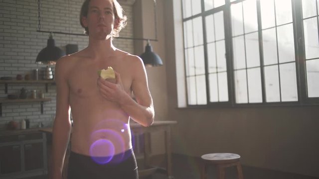 Portrait of a handsome man with a bare-chested eating an apple standing in the kitchen in the sunlight.
