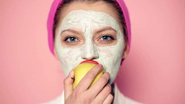 Close up of a girl with a mask on the face who eats an apple on a pink background. Concept of skin care and cleansing of the face. Concept of a healthy lifestyle.