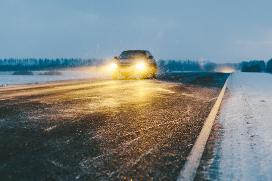 Car On Snowy Asphalt Road Drives In Dark Time With Bad Visibility. Out Of Focus Winter Landscape With Low Angle View