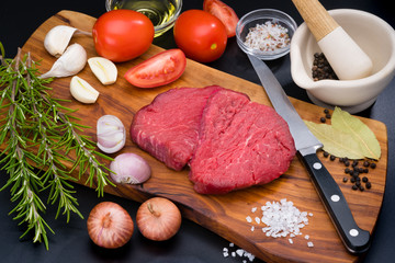 Two raw beef fillet steaks with spices and vegetables on a wooden board with a kitchen knife.