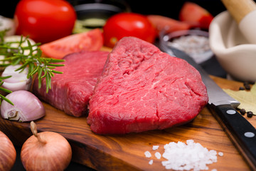 Two raw beef fillet steaks with spices and vegetables on a wooden board with a kitchen knife.