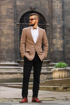 Fototapeta Young confident hipster man with beard in glasses posing on the street in old town. Attractive man wearing a suit, white shirt and light trousers.