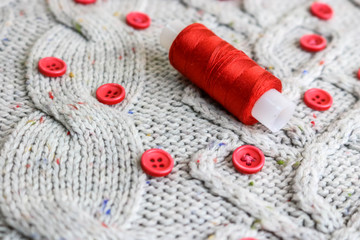 Beautiful texture of a soft warm natural sweater, fabrics with a knitted pattern of yarn and red small round buttons for sewing and a skein of red thread. The background