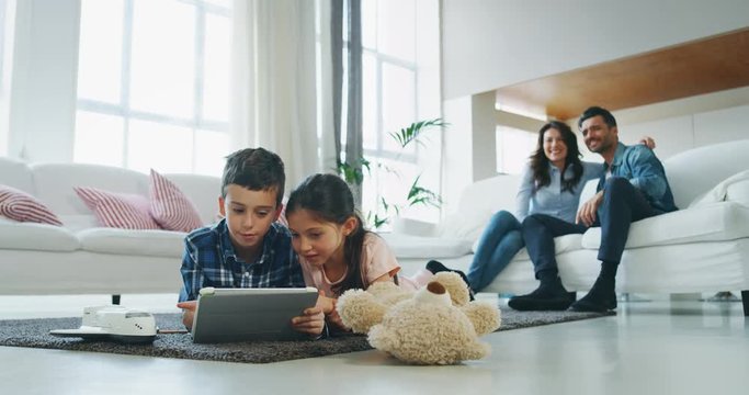 Portrait of happy kids using tablet having fun lying on the carpet in living room with their parents in slow motion. Shot with RED camera in 8K. Concept of  family entertainment, education, technology
