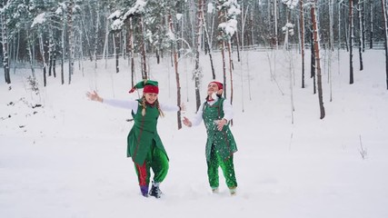 Funny elves in green suits with hats are throwing up snow and looking at it on the background of a winter park. Slow motion