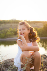 A small smiling girl with curly brown hair dressed white short dress sitting on the stones at the river on sunset time
