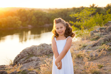 A small smiling laughing girl with curly brown hair dressed white short dress on the stones at the river on sunset time