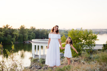 A small smiling laughing girl with curly brown hair dressed white short dress with young beautiful mother in long white dress on the stones at the river on sunset time