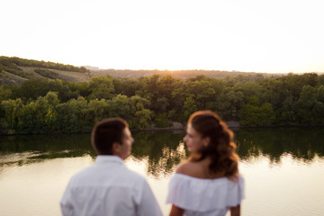 Young beautiful woman in long white dress with her husband on the stones at the river on sunset time