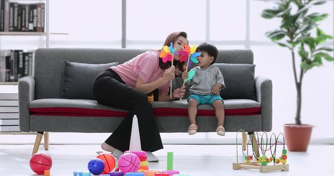 Asian mother sitting on sofa with her son, they playing and blowing colorful wind wheel toy, many toys on floor, concept for mother and son playing wind wheel toy in living room. 