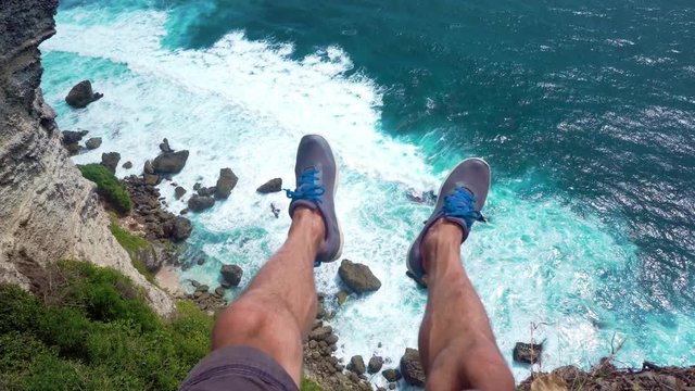first person view. The man traveler sits on the edge of the cliff with his legs dangling. At the bottom of the waves splashing and crashing on the rocks