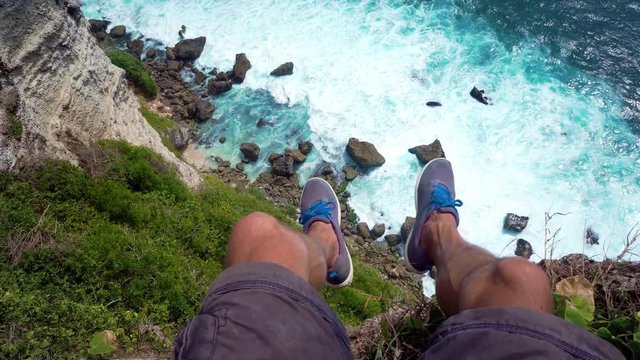 first person view. The man traveler sits on the edge of the cliff with his legs dangling. At the bottom of the waves splashing and crashing on the rocks