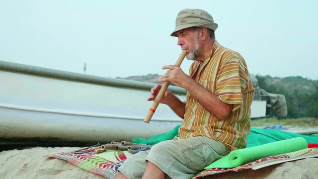 Senior man playing bamboo flute on the beach next to fishing boat