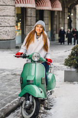 Vertical evening photo of beautiful young woman in warm clothes smiling while riding retro scooter in city street in cold winter snowy day.