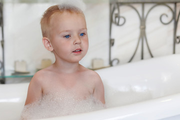 Cute caucasian baby boy with bright blue eyes and fair hair in the bathtube, with foam bubbles o the head and around. Indoors, copy space.