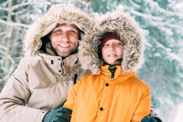 Father and son dressed in Warm Hooded Casual Parka Jacket Outerwear walking in snowy forest cheerful smiling faces portrait. Father and son relatives and winter outfit concept image. © Soloviova Liudmyla