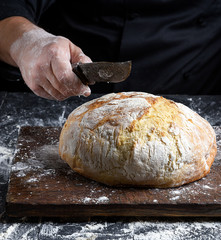 male chef hand holds knife over a whole round baked loaf of bread