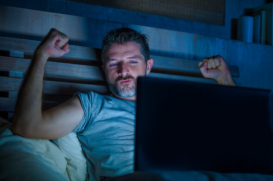man alone in bed playing cybersex using laptop computer watching porn sex movie late at night with lascivious pervert face expression in internet pornographic sexual content