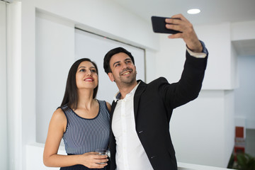 Smiling mixed race businessman photographing with colleague in office. Cheerful young business colleagues taking selfie in corridor. Social media concept