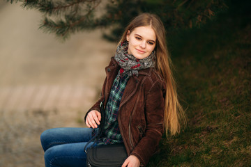 Attrective young girl sits by the embankment. Blond hair and brown jacket. Spring