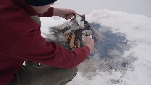 Traveling man pours hot coffee near camp fire an in winter time, surrounded by snow  near of the frozen lake. Concept adventure active vacations outdoor hiking sport