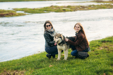 Naklejka na ściany i meble Two sisters walk with husky dog in front of river. Brunettle and blonde girl caress a dog