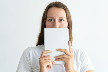 Young woman holding tablet computer in front of face. Pretty lady using digital device and looking at camera. Technology concept. Isolated front view on white background.