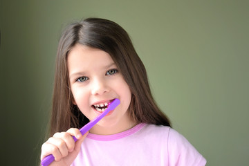 Long haired smiling beautiful caucasian white girl brushing her teeth with violet toothbrush on green background. Green eyed cute child in violet tshirt doing morning hygiene. Kid holding violet brush