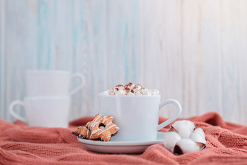 Coffee cup with marshmallow and stars cookies on warm knitted coral blanket, christmas lights and cotton flowers.