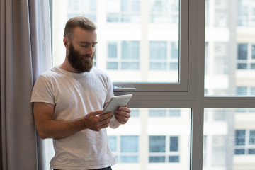 Serious handsome hipster guy using digital tablet at home. Pensive young bearded man standing at...