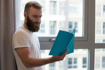 Serious hipster man in tshirt reading book. Concentrated young guy with beard standing at window. Hobby concept