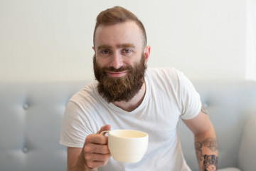Happy bearded young man in casual enjoying coffee break on couch. Stylish hipster drinking coffee at home. Weekend morning concept