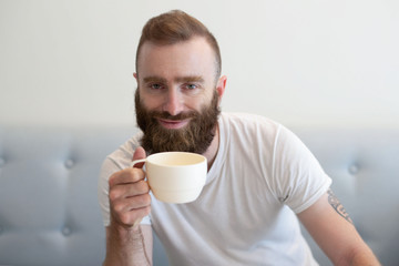 Peaceful hipster drinking coffee at home. Cheerful bearded young man in casual resting on couch and enjoying coffee break. Coffee break concept